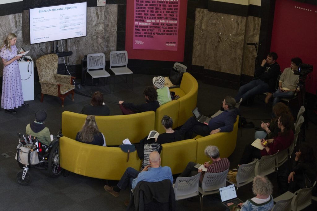 A woman presents a slideshow to a small audience seated on yellow couches and chairs. The room has a concrete wall with a pink poster and a mix of casual and attentive listeners, some with laptops and notebooks.