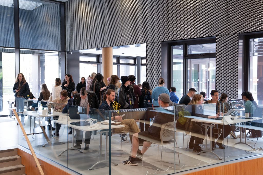 A busy university foyer with students in a line and others seated at desks, interacting with staff and guests. The modern setting features large windows and a dynamic design.