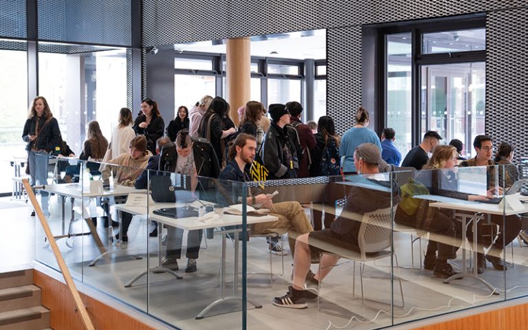 A busy university foyer with students in a line and others seated at desks, interacting with staff and guests. The modern setting features large windows and a dynamic design.