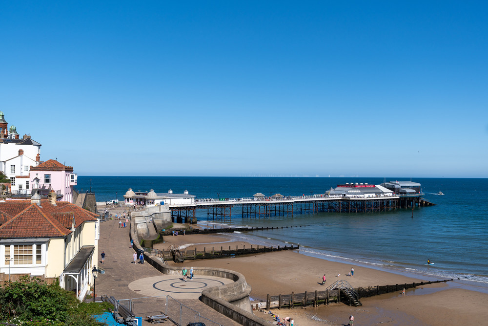 A coastal scene featuring a long pier extending over the water, a sandy beach with people walking, and buildings with red-tiled roofs on the left. The sky is clear and blue, and the sea appears calm.