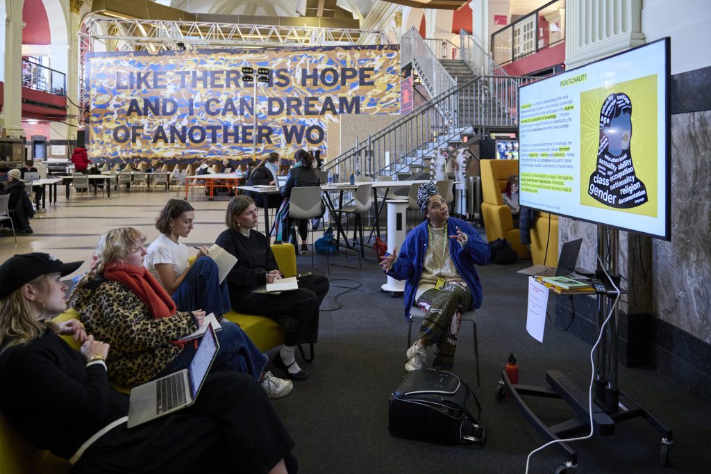 A group of people are seated in a casual setting, engaging with a speaker presenting from a laptop connected to a large screen. Behind them, a large artwork is displayed across the room.