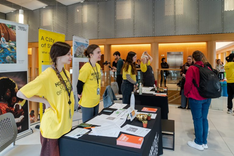 People are gathered in a well-lit indoor space with tables set up for an event. Several individuals in yellow shirts interact with visitors. Informational materials and flyers are spread across the tables. Banners and posters are in the background.