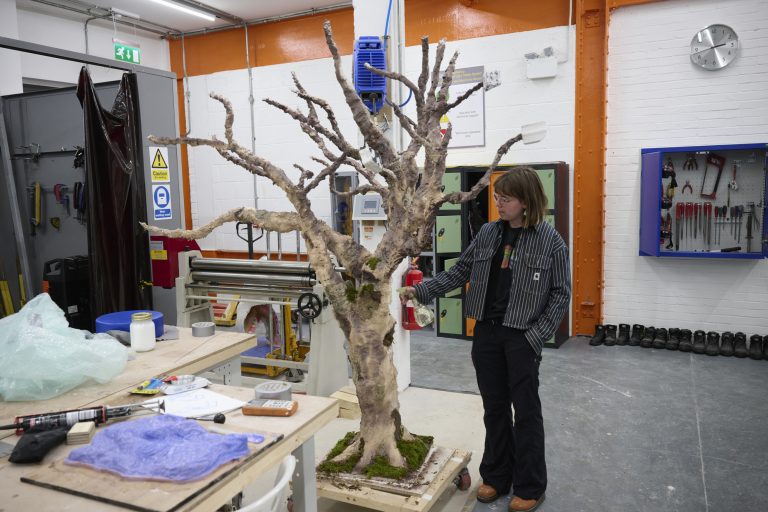 A person stands next to a large, realistic model of a bare tree in a workshop. The workshop is filled with various tools and equipment. The tree is mounted on a base, and the individual appears to be observing or working on it.