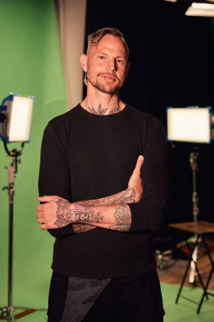 A person with tattoos on their arms and neck stands confidently with arms crossed in front of a green screen. They are wearing a black long-sleeve shirt and dark pants. Studio lights and equipment are visible in the background.