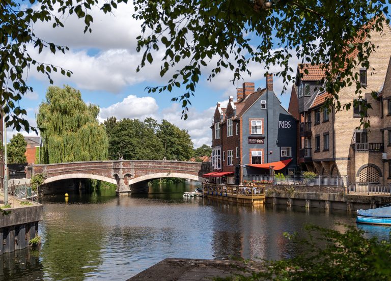 A serene riverside scene featuring a stone bridge over a calm river, flanked by quaint buildings, one of which houses a restaurant with outdoor seating under red umbrellas. Lush trees and leafy branches frame the image, while a few boats are tied up along the water's edge.