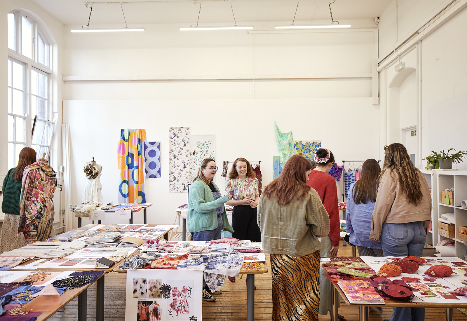 A group of people in a bright textile studio discussing the students work displayed on tables and walls.