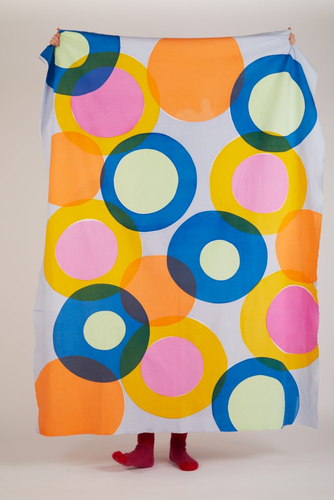 A person standing behind a large sheet or fabric with a pattern of overlapping colourful circles in various shades of orange, blue, green, pink, and yellow. Only the person's hands and feet, in red socks, are visible.