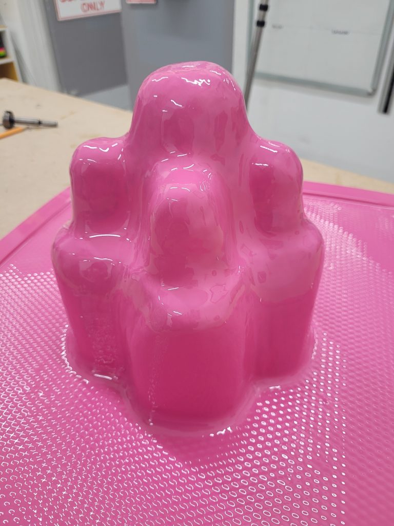 A glossy, pink, abstract sculpture with a smooth surface stands on a textured, pink base. The sculpture has multiple rounded, mound-like shapes of varying heights connected together. It is situated in an indoor space with industrial elements in the background.