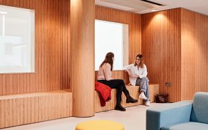 Two people are sitting and chatting in a modern room with wooden walls and accents. One is seated on a wooden bench with a red jacket beside them, while the other sits on a ledge. The room features a yellow round ottoman, a blue chair, and a handbag on the floor.