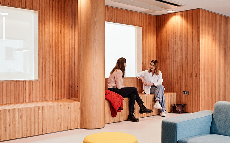 Two people are sitting and chatting in a modern room with wooden walls and accents. One is seated on a wooden bench with a red jacket beside them, while the other sits on a ledge. The room features a yellow round ottoman, a blue chair, and a handbag on the floor.