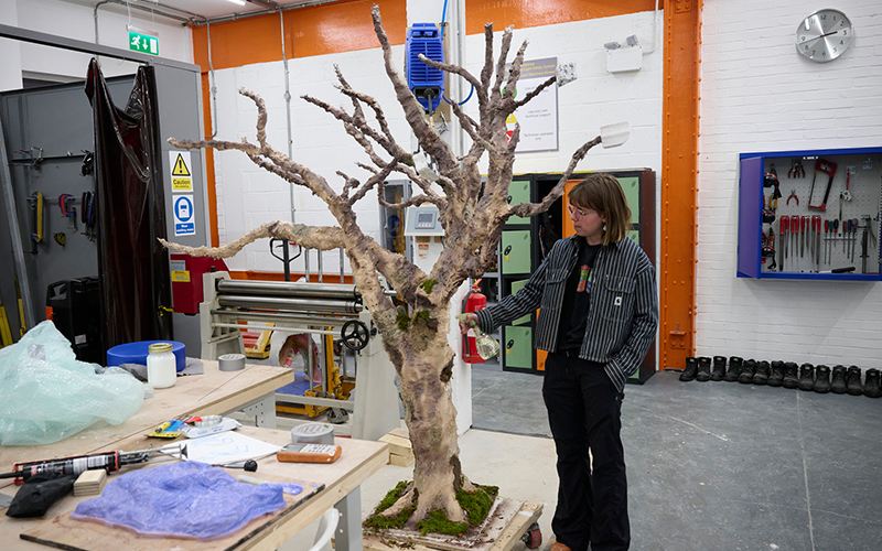 A person stands next to a large, realistic model of a bare tree in a workshop. The workshop is filled with various tools and equipment. The tree is mounted on a base, and the individual appears to be observing or working on it.