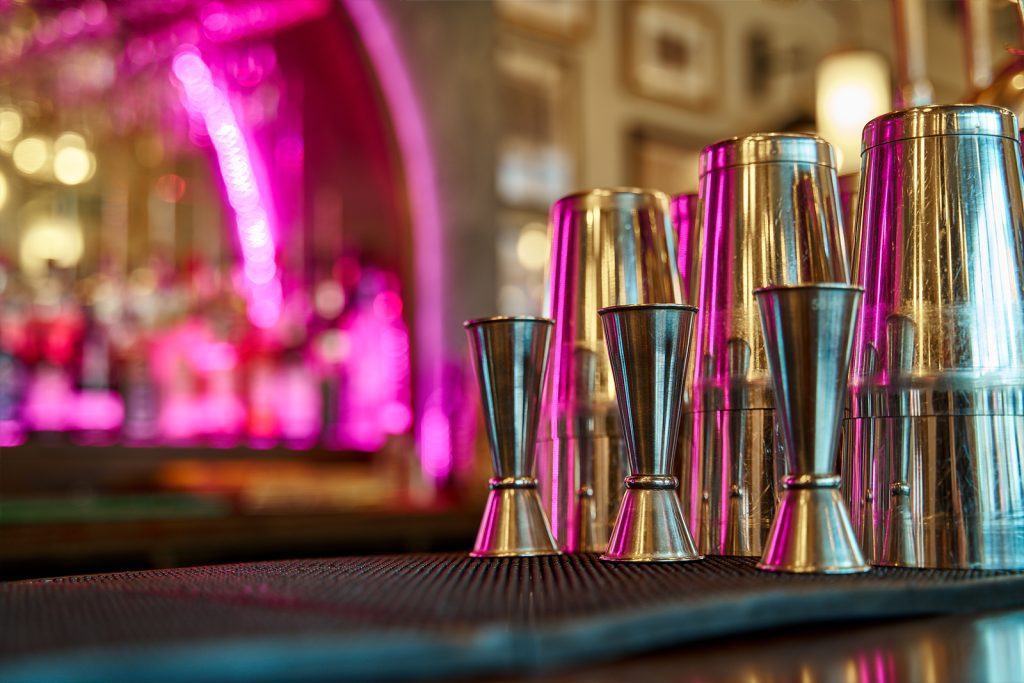 A row of shiny metal cocktail shakers and jiggers arranged on a black mat on a bar counter. The background is out of focus, showcasing a brightly lit bar with purple neon lights and various bottles.