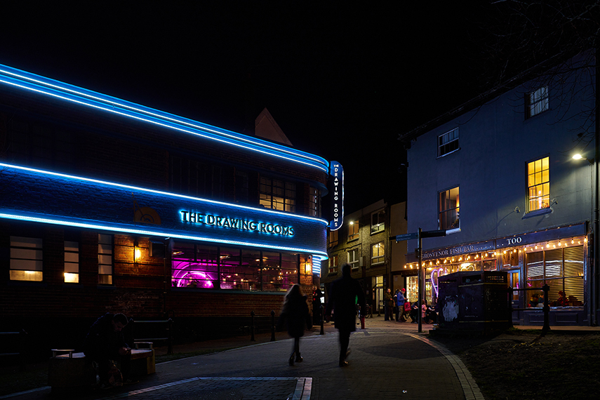 A nighttime street scene with a brightly lit building on the left featuring neon blue lights and a sign that reads "The Drawing Rooms." On the right, people walk past a warmly lit café or restaurant. The street is paved and lined with buildings.