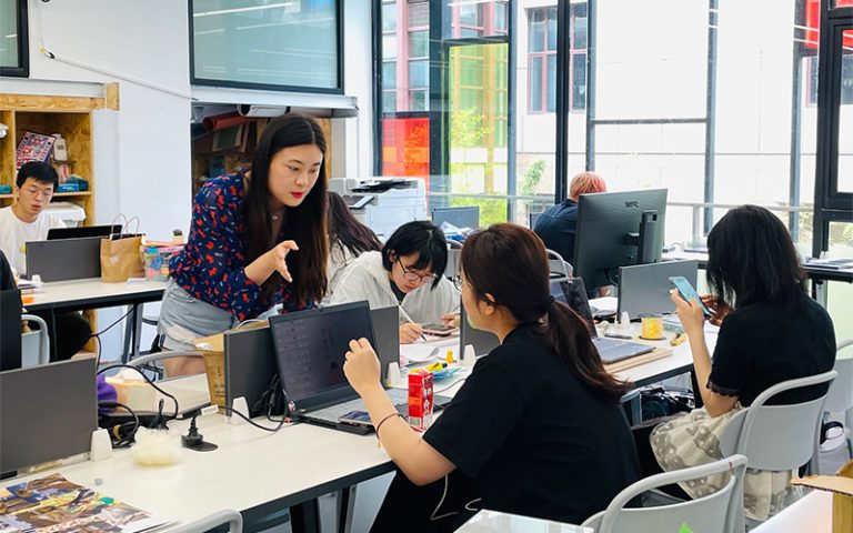 A group of people work in a brightly lit office with large windows. A woman in a blue floral blouse talks to another woman seated at a desk with a laptop. Other colleagues are seated at desks, using laptops and smartphones, creating a collaborative atmosphere.