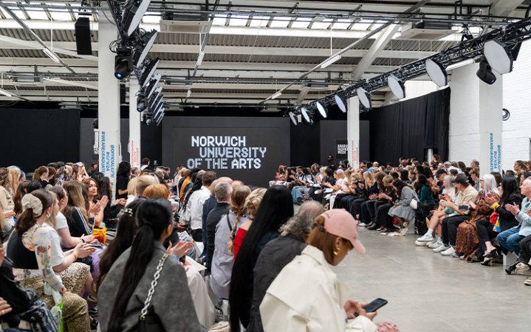 A crowded indoor venue with people sitting on both sides of a runway, watching a fashion event. At the end of the runway, there is a large screen displaying "Norwich University of the Arts." The audience is engaged, with some clapping and others taking photos.