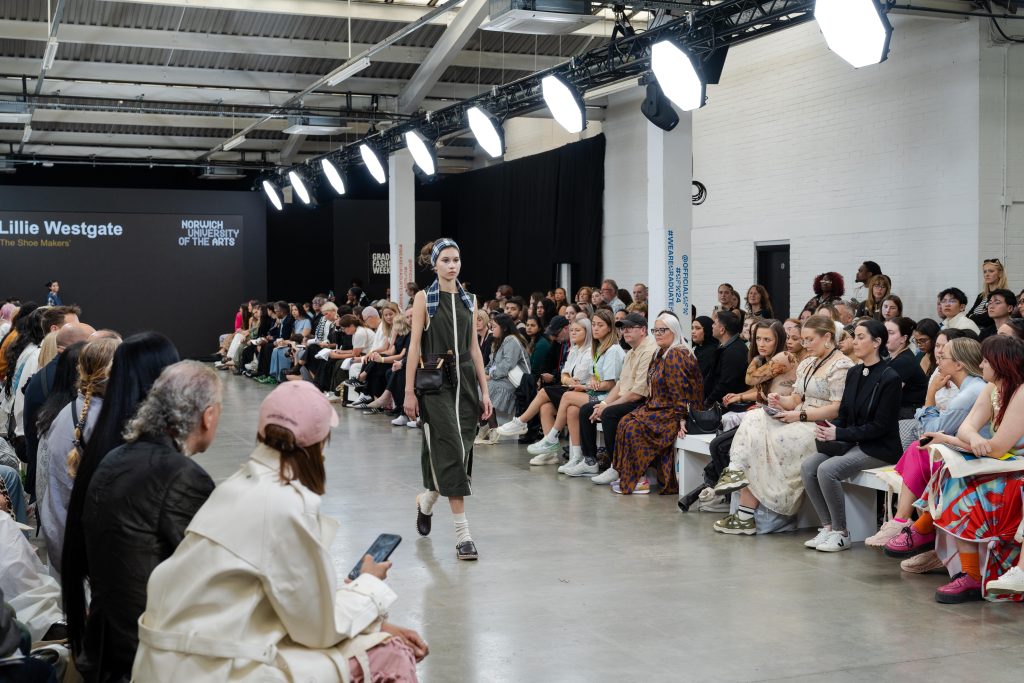 A fashion model walks down the runway wearing a dark green and white outfit with black boots and a headband. The audience, seated on both sides, watches attentively. There's a display screen in the background with the text Lillie Westgate.