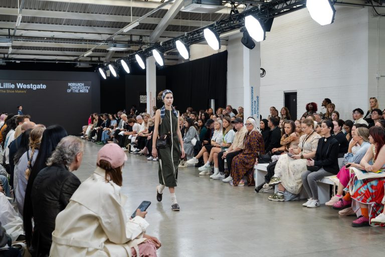 A fashion model walks down the runway wearing a dark green and white outfit with black boots and a headband. The audience, seated on both sides, watches attentively. There's a display screen in the background with the text Lillie Westgate.