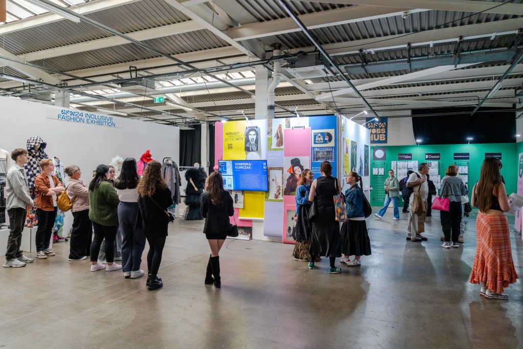 A group of people are gathered in an indoor space, observing exhibits and displays on various topics. Several walls feature colourful posters and information boards. The room has a high ceiling with industrial-style lighting and beams.