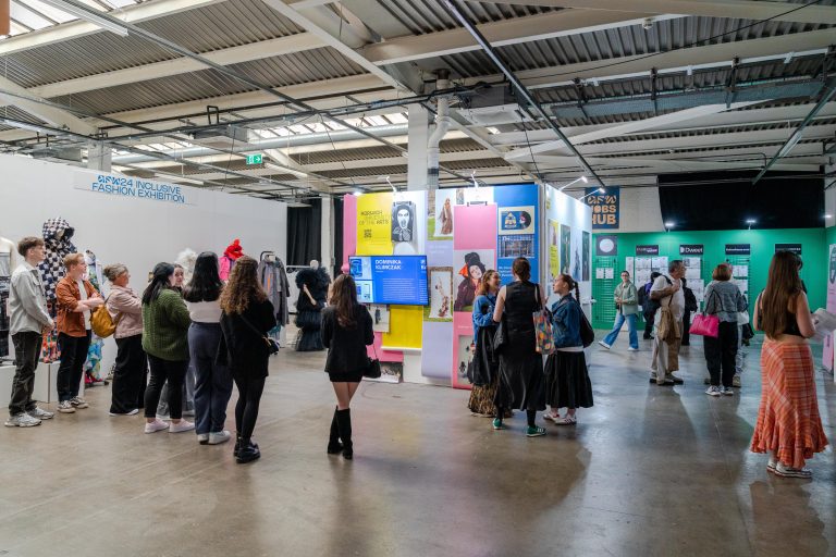 A group of people are gathered in an indoor space, observing exhibits and displays on various topics. Several walls feature colorful posters and information boards. The room has a high ceiling with industrial-style lighting and beams.