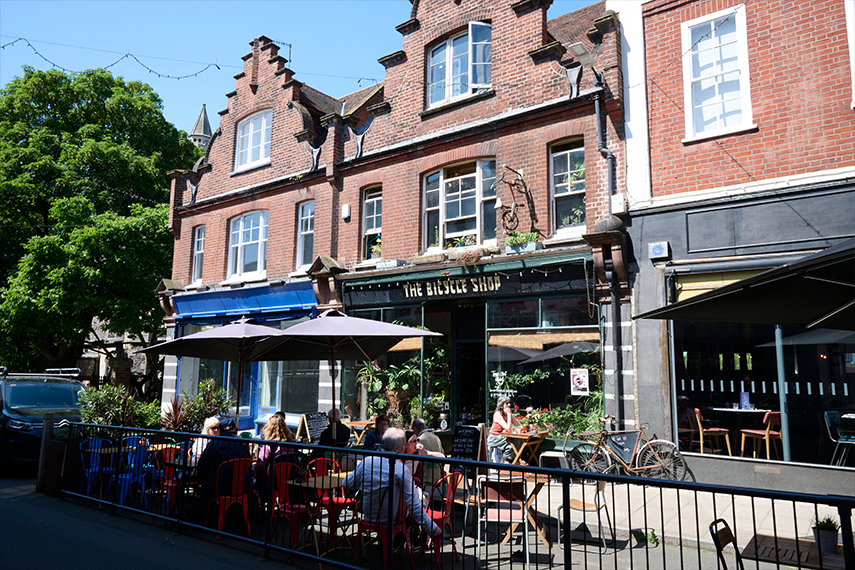 A lively street scene featuring a group of people sitting at outdoor tables under umbrellas in front of a building with brick facades and tall windows. A hanging sign reads "The Bicycle Shop," and bicycles are parked by the entrance. Sunlight bathes the area.