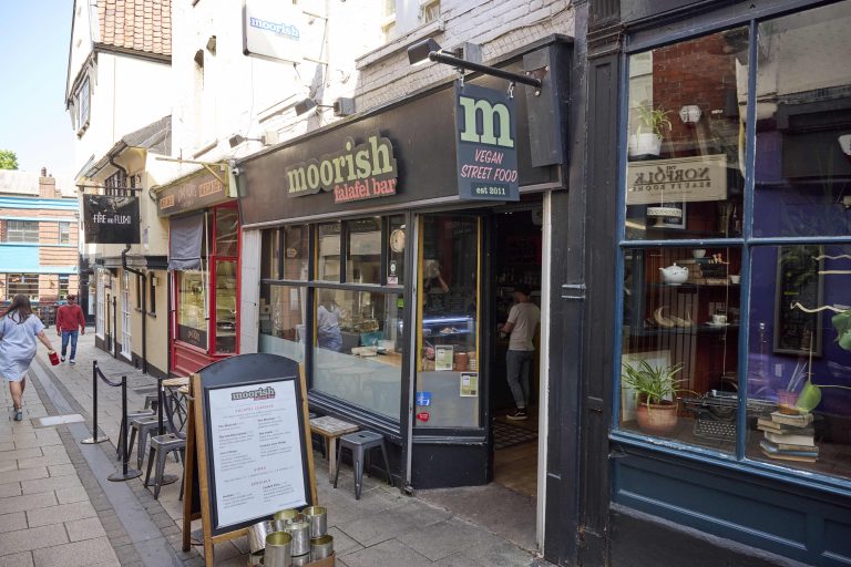 A street view of a small eatery named "Moorish Falafel Bar" offering vegan street food. The storefront has a black sign with white and green letters. A sandwich board menu is placed outside next to chairs and tables.