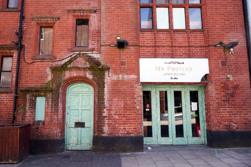 A red brick building with two green doors. The right side has a sign reading "Mr Postles' Apothecary" above a door with large glass panels. The left side is a wooden door under an arched stone frame with a small rectangular window above.