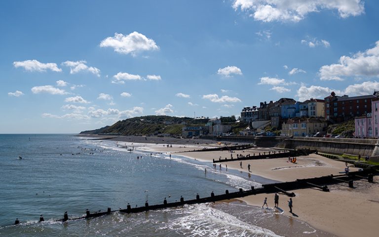 A coastal townscape with a beach featuring people walking along the shore and wooden groynes extending into the sea. The calm water reflects the sunny, partly cloudy sky. Buildings with colourful facades line the background along the coastline.