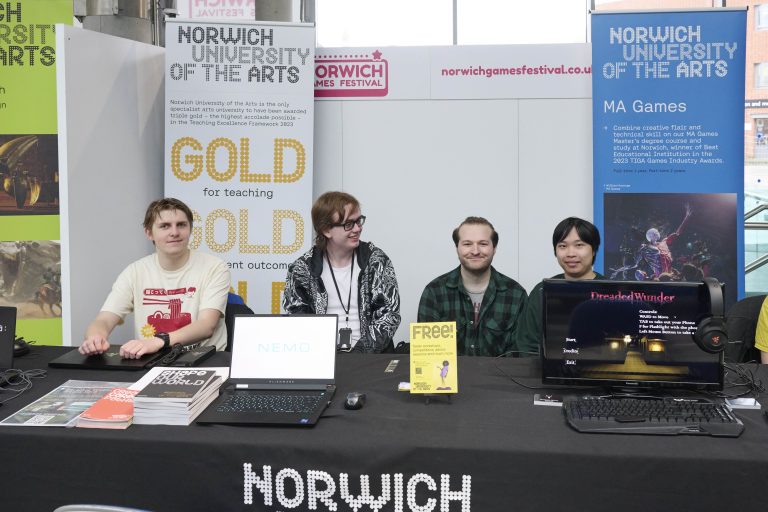 A group of four people sit behind a table displaying laptops and promotional materials at the Norwich University of the Arts booth. Banners in the background advertise the university's MA Games program and its high teaching standards.