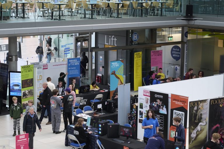 People attending a technology and gaming event in a modern indoor venue with multiple booths showcasing various products and information. Several attendees are engaging with exhibits, while others are walking around the space. Bright banners and posters are visible.