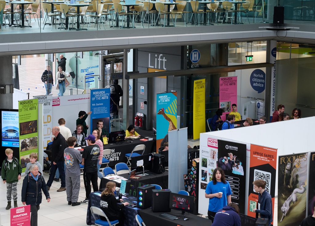 People attending a technology and gaming event in a modern indoor venue with multiple booths showcasing various products and information. Several attendees are engaging with exhibits, while others are walking around the space. Bright banners and posters are visible.