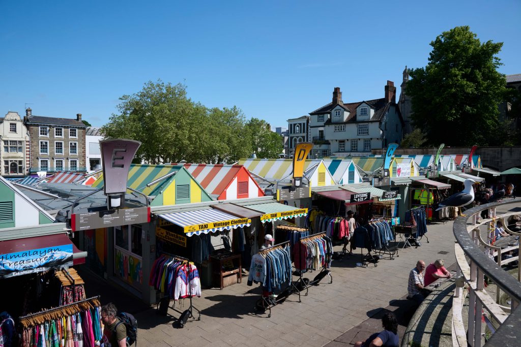 A vibrant outdoor market with colourful striped stalls. Shoppers browse through hanging clothes while others sit on steps to the right. Trees and historic buildings are visible in the background under a clear blue sky.