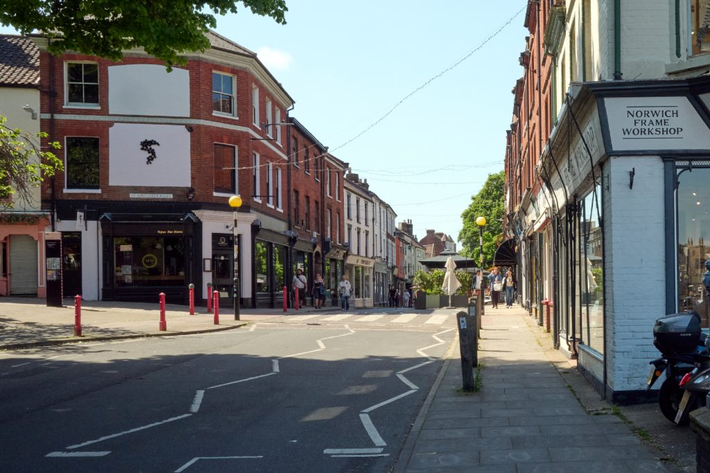 A quiet street scene with pastel-coloured buildings. There are trees, red bollards, and a clear sky.