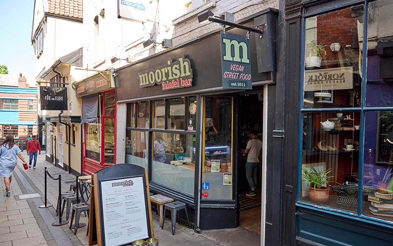 A street view of a small eatery named "Moorish Falafel Bar" offering vegan street food. The storefront has a black sign with white and green letters. A sandwich board menu is placed outside next to chairs and tables.