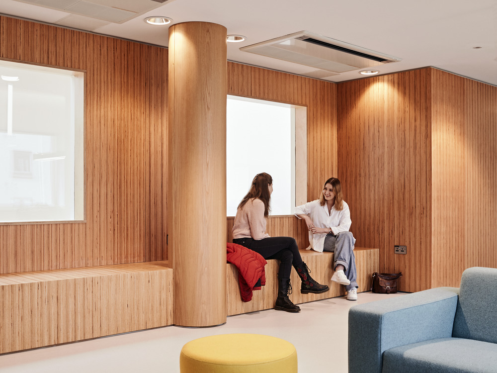 Two people are sitting and chatting in a modern room with wooden walls and accents. One is seated on a wooden bench with a red jacket beside them, while the other sits on a ledge. The room features a yellow round ottoman, a blue chair, and a handbag on the floor.