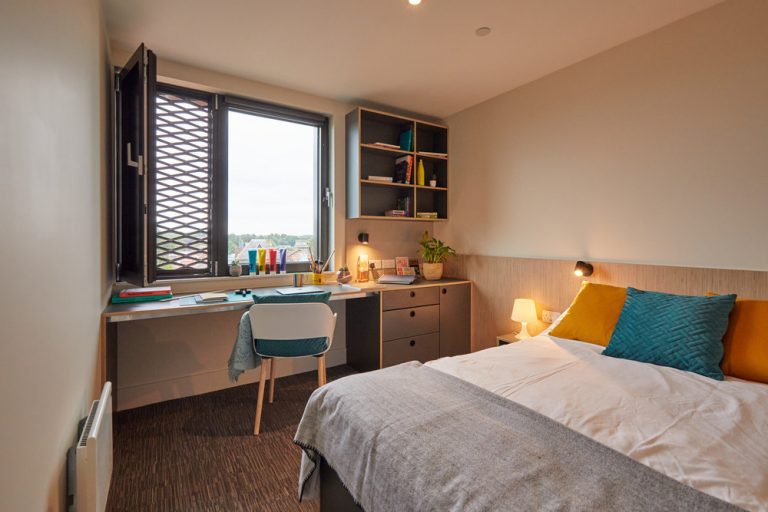 A tidy bedroom features a neatly made bed with gray and mustard pillows, a desk with stationery, books, a lamp, and a desktop underneath a window with an open pane. There's a plant on the desk, and shelves with books and decor elements on the wall.