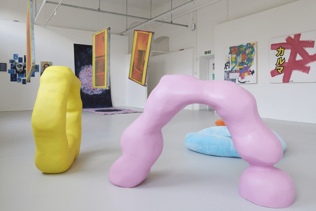 A contemporary art gallery showcases colorful abstract sculptures—including a yellow upright form—created by an artist with an MA Fine Art degree, surrounded by vibrant paintings, hanging fabric pieces, and a blue cushion on a white floor.