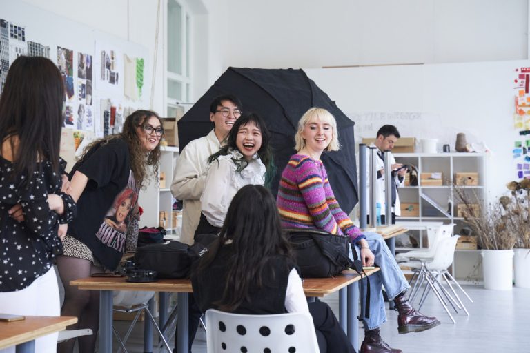 A group of five young adults are gathered around a desk in an art studio, laughing and smiling. The studio is decorated with colorful artwork on the walls, and a softbox lighting setup is visible in the background. One person sits on a desk, facing the group.