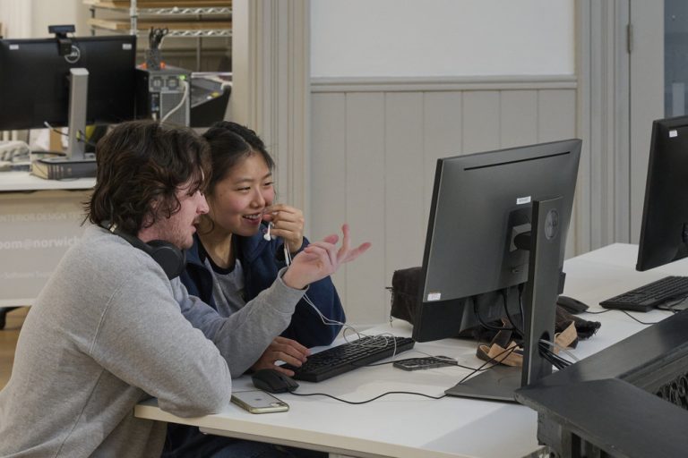 Two people seated at a desk, focused on a computer screen. One, with long hair and wearing headphones around his neck, gestures with a pen. The other, a woman with her hair tied back, smiles while pointing at the monitor. Several other computers and equipment are in the background.