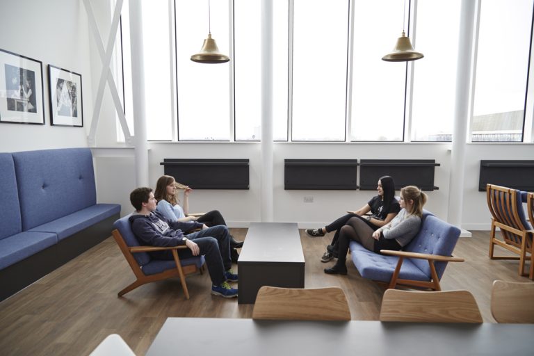 Four people are seated and engaged in conversation in a modern, well-lit room. The space features large windows, blue sofas, wooden chairs, and hanging pendant lights. The decor includes framed photos on white walls and a central dark coffee table.