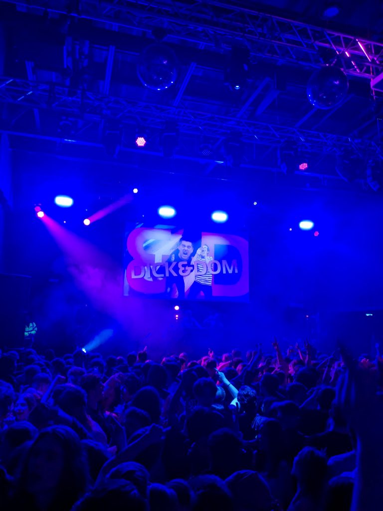 A vibrant concert scene with a large crowd of people dancing under blue and purple lights. On the stage, there's a large screen displaying the text "DICK & DOM" along with images of two individuals. The atmosphere is energetic and filled with excitement.