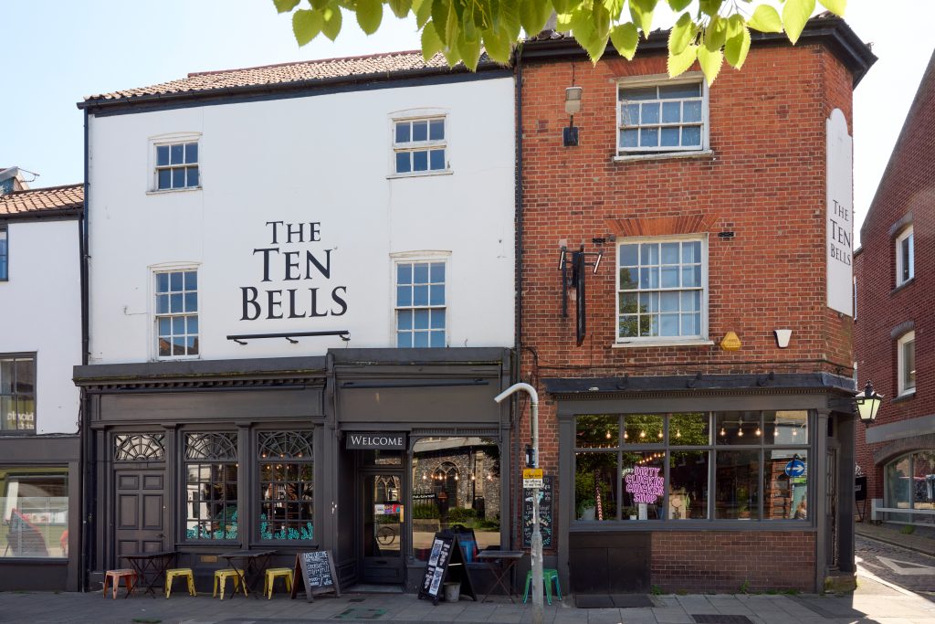 A street view of The Ten Bells pub, housed in a historic brick building with white upper floors. The pub has large windows, a black sign with its name, and colourful stools outside the entrance. There are trees and neighboring buildings visible in the background.