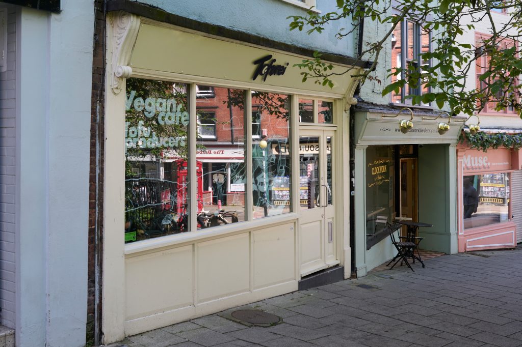 Street view of a row of shops, with a light-colored storefront named "Tofurei" displaying "Vegan cafe" on the window. More shops are visible in the background.