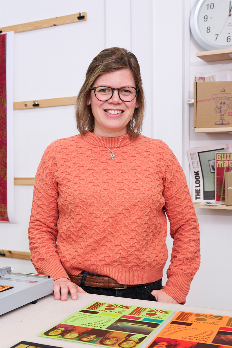 A woman wearing glasses and an orange textured sweater stands smiling behind a desk with colorful papers and books; a clock and shelves with books are visible in the background.