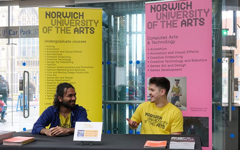 Two people in yellow "Norwich University of the Arts" shirts sit behind a table with brochures, smiling and talking. They are in front of two banners advertising courses offered by the university. One banner is pink, and the other is yellow.