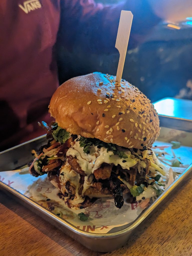 Close-up of a gourmet burger in a tray. The burger has a sesame seed bun and is filled with various ingredients. A wooden skewer holds the burger together.