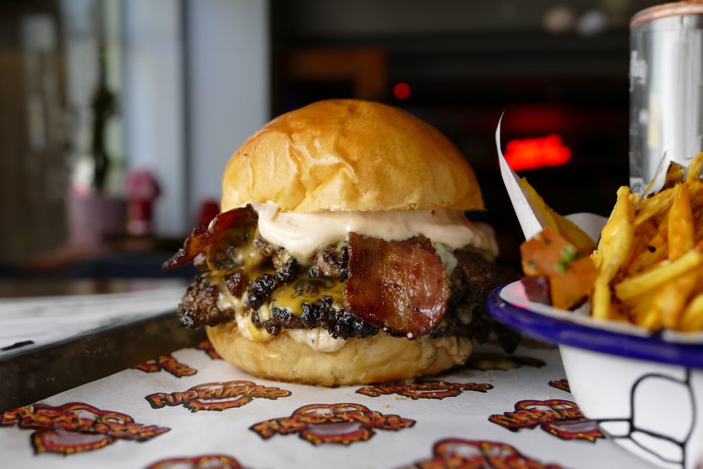 A juicy burger topped with bacon on a toasted bun is placed on a tray lined with paper featuring playful designs. In the background, a bowl of seasoned fries is partially visible. The setting appears to be a casual dining restaurant.