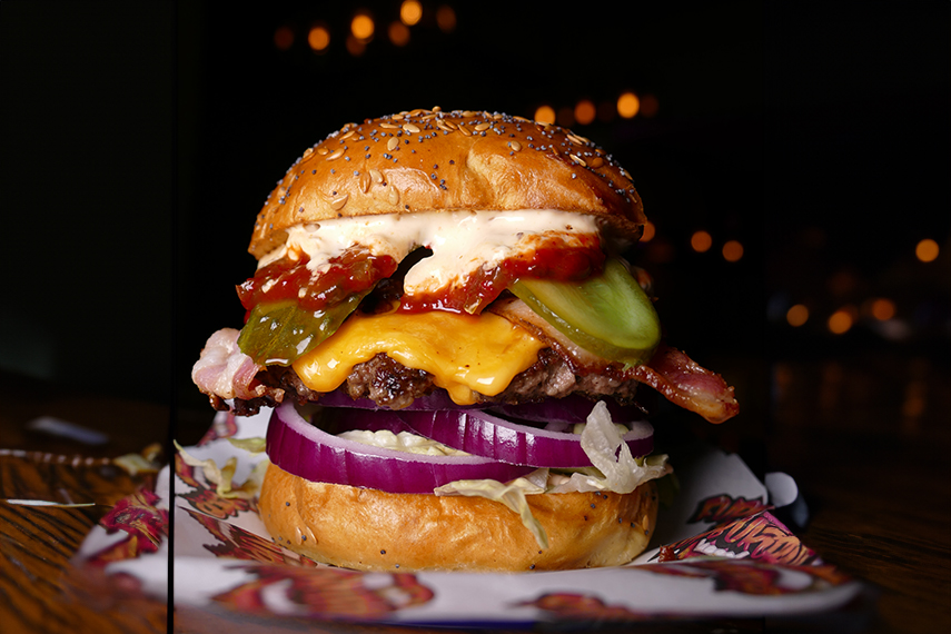 A juicy burger with a sesame seed bun is filled with melted cheese, crispy bacon, pickles, onions, lettuce, and a generous serving of sauce. The burger is set against a dark, blurred background, enhancing its vibrant colors and appetizing appearance.