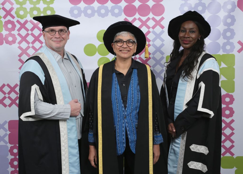 Three people in academic regalia smile at the camera standing in front of a colourful geometric background. They wear black gowns with light blue trim and black hats. The person in the center has an additional blue and gold robe.