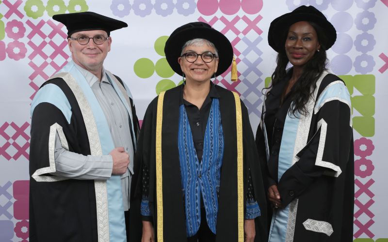 Three people wearing academic robes and hats stand together against a backdrop decorated with colourful geometric patterns. The person in the middle is wearing a robe with a gold trim, while the other two wear robes with light blue trims. All are smiling.