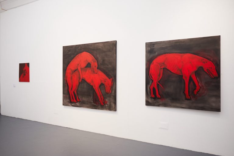 Three paintings on a white wall depict red dogs on dark backgrounds. The right painting shows a single dog standing. The middle painting shows two dogs engaged with each other. The left, smaller painting also features one dog in partial view. The setting is minimal and modern.